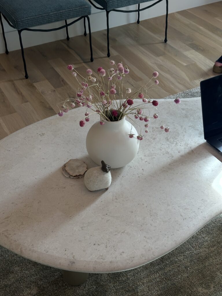 Organic-shaped stone coffee table styled with a white matte vase of dried pink flowers, natural stone coasters, and a small sculptural object, set on a soft rug with wood flooring and patterned chairs in the background.