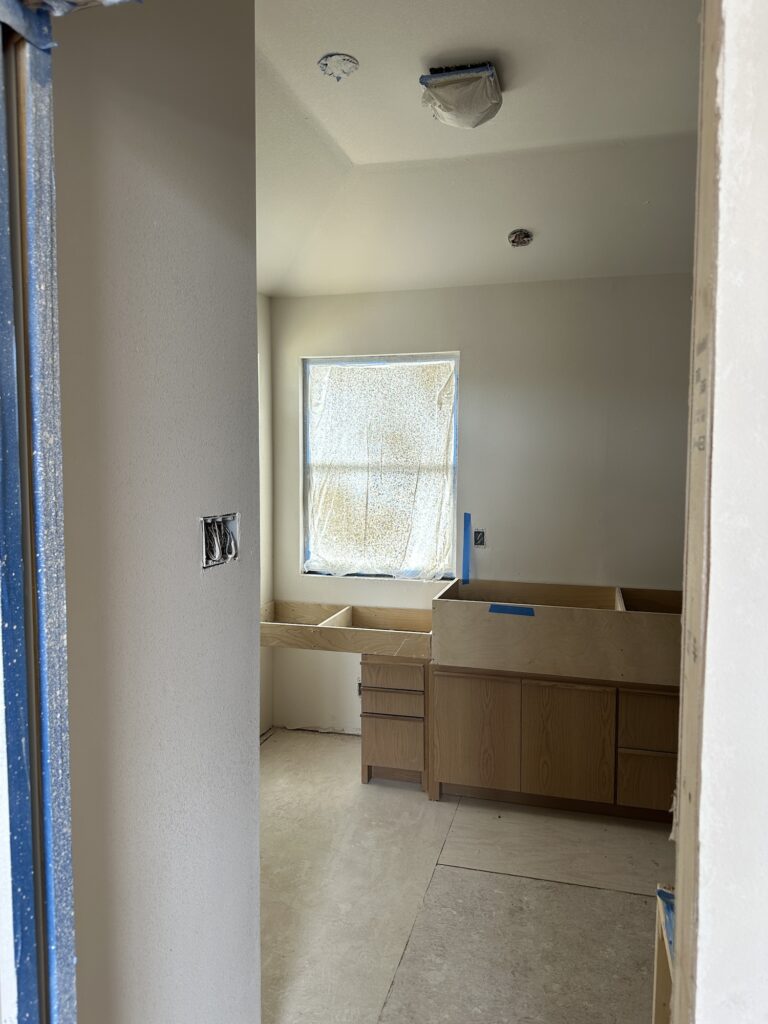 Primary bathroom under construction at Casa Urbana, showing custom white oak cabinetry installation beneath a window with natural light filtered through protective covering. Drywall and ceiling prep are in progress, with flooring and fixtures yet to be installed.
