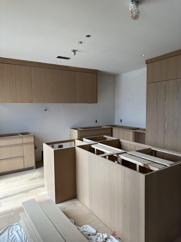 In-progress view of a custom kitchen renovation featuring natural white oak cabinetry with flat-panel doors and a large center island. Upper and base cabinets are installed, with countertops and appliances yet to be added. Light wood flooring and recessed lighting create a clean, modern foundation.